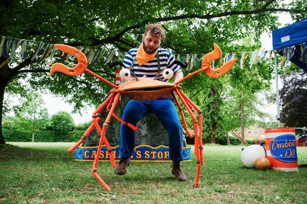 A man with brown hair dressed in blue and white operates an orange crab puppet standing on grass and in front of trees.