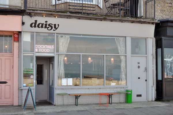 A photograph of a bar with a grey front, with a bench, tables and rubbish bin outside. The name of the bar in Daisy. There is a neon sign above the door reading 'cocktails and food'.
