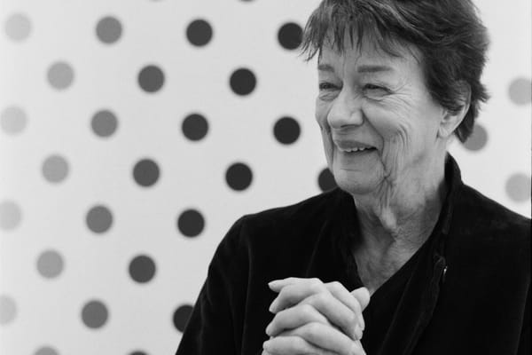 A black and white photo of an elderly woman dressed in black, smiling and clasping her hands together against a backdrop of polka dots.